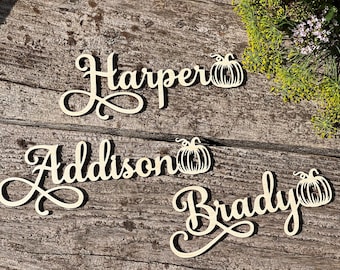 May include: Wooden name cutouts with decorative pumpkin accents. The names "Harper", "Addison", and "Brady" are displayed in a cursive font. The cutouts are light-colored and placed on a rustic wooden surface.
