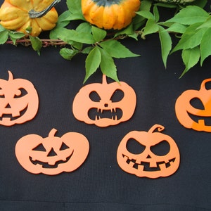 Può includere: Cinque ritagli di zucche arancioni con facce intagliate, perfetti per le decorazioni di Halloween. Le zucche sono disposte su una superficie nera, con foglie verdi e piccole zucche sullo sfondo.