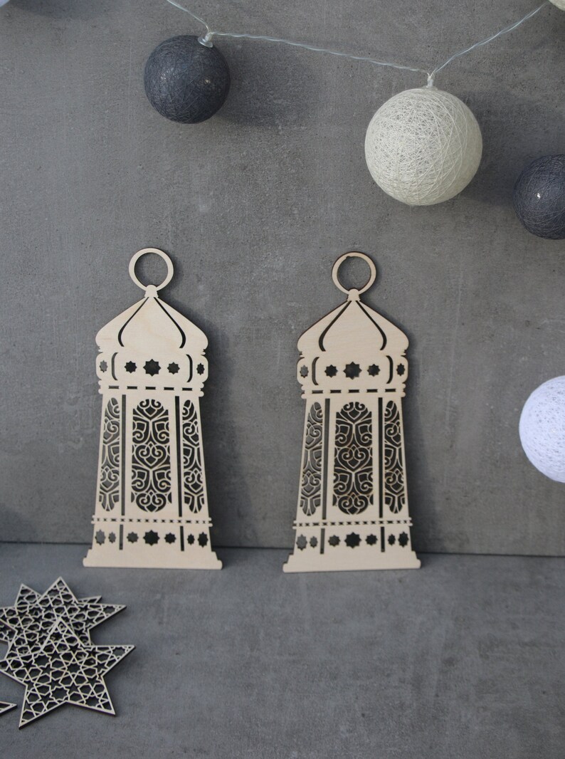 May include: Two wooden laser cut lanterns with intricate designs. The lanterns are light brown and have a white background. The lanterns are hanging from a string of lights.
