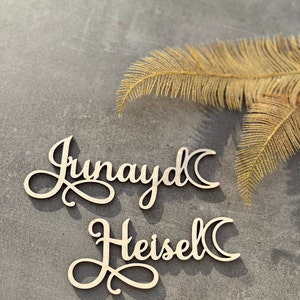 Op de afbeelding: Twee houten naamuitsparingen, "Junayd" en "Heisel", in een cursief lettertype, elk met een maansikkelontwerp. De namen zijn lichtgekleurd tegen een grijze achtergrond, met een gouden veer rechtsboven.