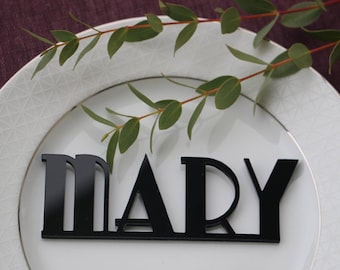 May include: A white plate with a gold rim and a geometric pattern. A black acrylic name card with the name "MARY" is placed on the plate. A sprig of green eucalyptus is placed on the plate.