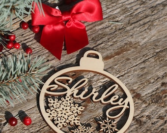 May include: A wooden Christmas ornament shaped like a bauble with the name "Shirley" cut out in a decorative font. The ornament is decorated with snowflakes and is surrounded by red berries and a green pine branch.