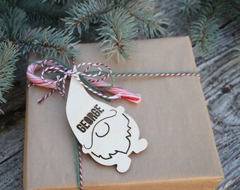 Puede incluir: Una bolsa de regalo de papel marrón con un bastón de caramelo rojo y blanco y un adorno de gnomo de madera con el nombre "George" en él.