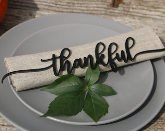Pode incluir: Um porta-guardanapo de madeira preto com a palavra "thankful" em uma fonte cursiva. O porta-guardanapo está colocado em um guardanapo cinza, que está em um prato cinza. Uma folha verde está colocada no prato.