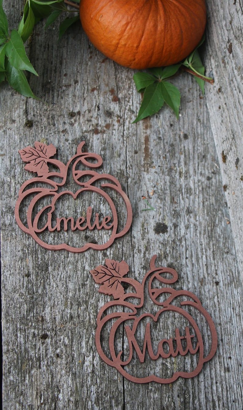 Puede incluir: Dos tarjetas de lugar de madera en forma de calabaza con los nombres "Ameli&eacute;" y "Matt" recortados. Las tarjetas de lugar est&aacute;n sobre una superficie de madera r&uacute;stica.