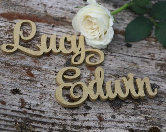 May include: Two wooden signs with the names "Lucy" and "Edwin" in cursive script, placed on a rustic wooden surface with a white rose.