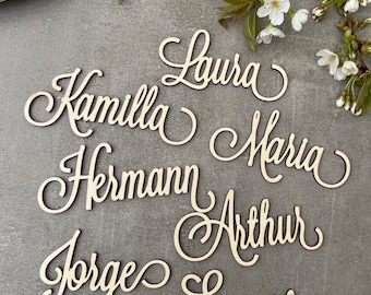 May include: Wooden name cutouts in a cursive font, including names like Laura, Kamilla, Maria, and Arthur. The names are arranged on a gray surface, with a sprig of white flowers in the background. Ideal for wedding or event decor.