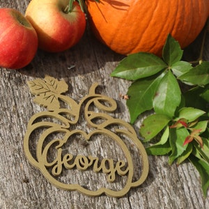 Puede incluir: Un adorno de madera con forma de calabaza con el nombre "George" recortado en el centro. El adorno es marr&oacute;n y tiene un dise&ntilde;o de hoja en la parte superior. El adorno est&aacute; sobre una superficie de madera con calabazas y manzanas en el fondo.