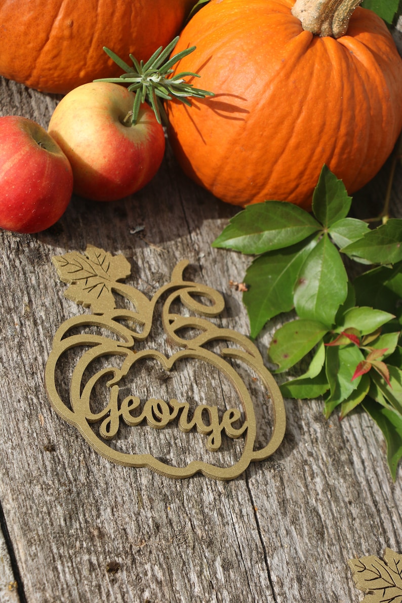 Puede incluir: Un adorno de madera con forma de calabaza con el nombre "George" recortado en el centro. El adorno es marr&oacute;n y tiene un dise&ntilde;o de hoja en la parte superior. El adorno est&aacute; sobre una superficie de madera con calabazas y manzanas en el fondo.