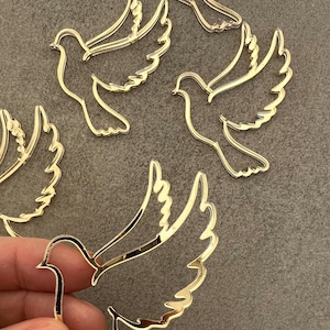 Può includere: Ritagli di colombe in metallo dorato con ali spiegate. Diverse forme di colombe sono sparse su una superficie grigia. Un ritaglio è tenuto in mano. I ritagli di colombe sono probabilmente per scopi artigianali o decorativi.