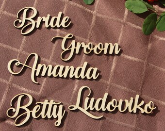 May include: Wooden name place cards with the words "Bride", "Groom", "Amanda", "Betty Ludovko", and "Teddy" written in cursive script. The cards are arranged on a brown fabric background.