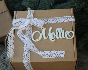 Puede incluir: Una caja de regalo de cartón marrón con una cinta de encaje blanco y una etiqueta de madera blanca que dice "Mollie".