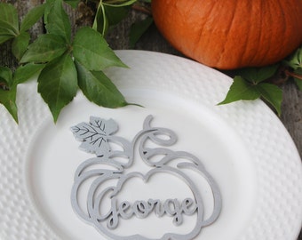 Puede incluir: Un plato blanco con borde texturizado sostiene una tarjeta de lugar plateada en forma de calabaza que dice "George". Una calabaza naranja y hojas verdes en el fondo sugieren un tema de otoño o Acción de Gracias.