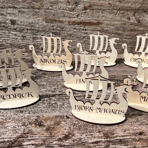 Op de afbeelding: Houten Viking schip naamkaarten met namen zoals Fredrick en Marie. De schepen hebben gedetailleerde uitsparingen van zeilen en rompen. De namen zijn in een decoratief lettertype, ideaal voor een thema-evenement.