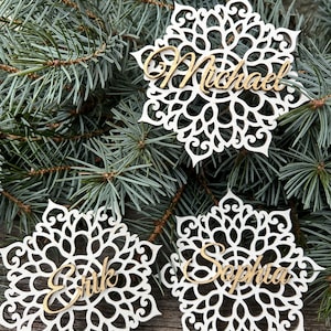 May include: Three white snowflake-shaped place cards with laser-cut designs. Each card has a gold-coloured name: Michael, Erik, and Sophia. The cards rest on a wooden surface with evergreen branches in the background.