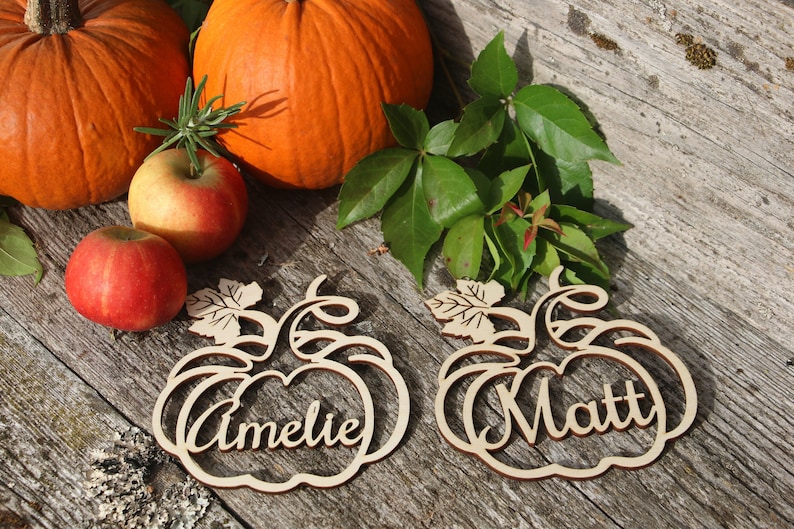 Puede incluir: Dos porta tarjetas de madera en forma de calabaza con los nombres "Amelie" y "Matt" escritos en ellos. Los porta tarjetas est&aacute;n sobre una superficie de madera con dos calabazas y manzanas.