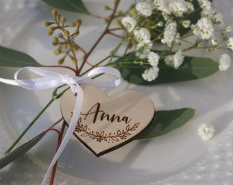 May include: A wooden heart-shaped place card with the name "Anna" engraved on it. The heart is tied with a white ribbon and decorated with a floral design.