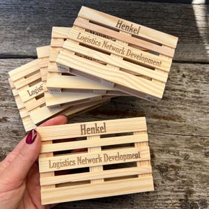 May include: A stack of small, light-colored wooden pallet-shaped coasters. The coasters have the name "Henkel" and "Logistics Network Development" engraved on them. One coaster is held by a person with a maroon nail polish.