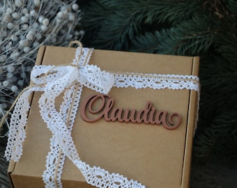 Puede incluir: Una caja de regalo marrón con una cinta de encaje blanco y una etiqueta de madera con el nombre "Claudia".