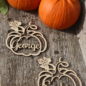 Puede incluir: Dos tarjetas de lugar de madera en forma de calabaza con los nombres "George" y "Sophia" escritos en cursiva. Las tarjetas de lugar est&aacute;n sobre una superficie de madera con dos calabazas naranjas.