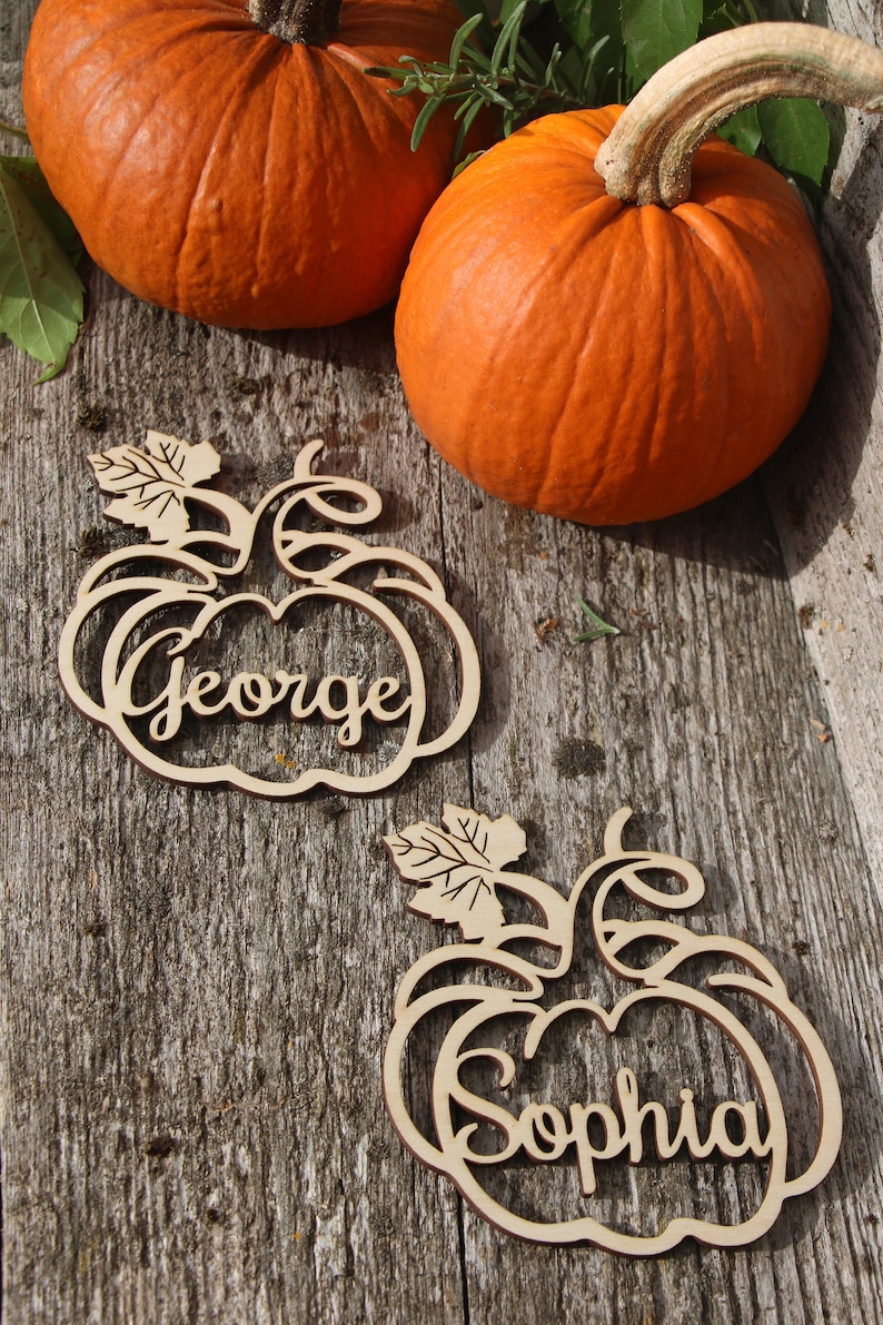 Puede incluir: Dos tarjetas de lugar de madera en forma de calabaza con los nombres "George" y "Sophia" escritos en cursiva. Las tarjetas de lugar est&aacute;n sobre una superficie de madera con dos calabazas naranjas.