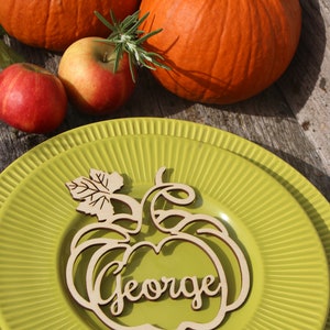 Puede incluir: Una tarjeta de lugar de madera en forma de calabaza con el nombre "George" tallado en ella. La tarjeta de lugar est&aacute; sobre un plato verde con un borde decorativo. Hay dos calabazas y manzanas en el fondo.