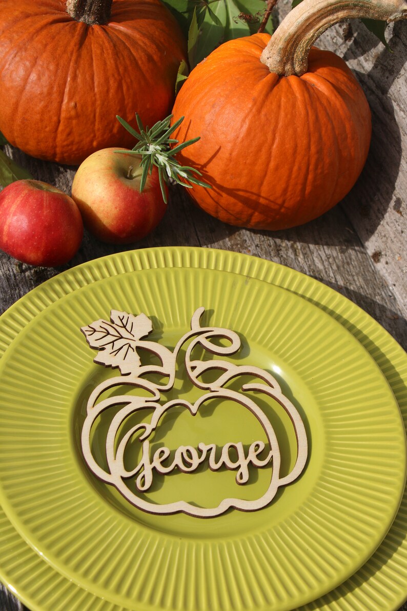 Puede incluir: Una tarjeta de lugar de madera en forma de calabaza con el nombre "George" tallado en ella. La tarjeta de lugar est&aacute; sobre un plato verde con un borde decorativo. Hay dos calabazas y manzanas en el fondo.