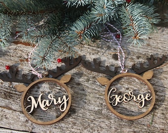 May include: Two wooden reindeer ornaments with the names "Mary" and "George" cut out in the center. The ornaments are hanging from twine and are against a rustic wooden background.