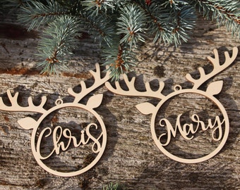 May include: Two wooden Christmas ornaments in the shape of reindeer heads, each with a circular center. One ornament says "Chris" and the other says "Mary" in cursive script. The ornaments are on a wooden surface with evergreen branches.