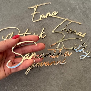 Può includere: Ritagli di nomi in acrilico dorato in un font corsivo. I nomi includono Emma, Patrick, Thomas, Kathy, Samantha e Giovanni. Il nome Samantha è tenuto in mano con smalto rosso.