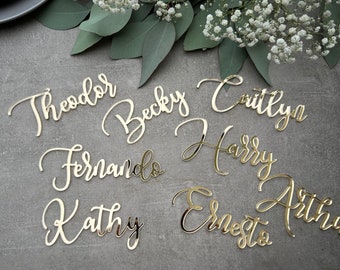 May include: Gold mirror acrylic place cards with the names Theodore, Becky, Coutlyn, Harry, Fernando, Kathy, Ernest, and Arthur written in cursive.