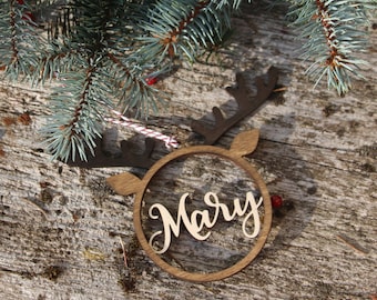 May include: A wooden Christmas ornament in the shape of a reindeer head with the name "Mary" in a cursive font. The ornament is brown and has a string for hanging. The background is a rustic wooden surface with evergreen branches.