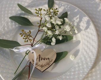 May include: A wooden heart-shaped place card with the name "Anna" engraved on it. The place card is tied to a sprig of greenery with white baby's breath and a white ribbon.