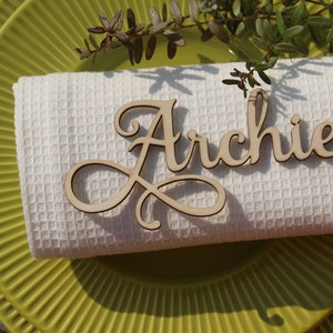 Puede incluir: Una servilleta de tela blanca con una tarjeta de nombre de madera que dice "Archie" en escritura cursiva. La servilleta est&aacute; encima de un plato verde.