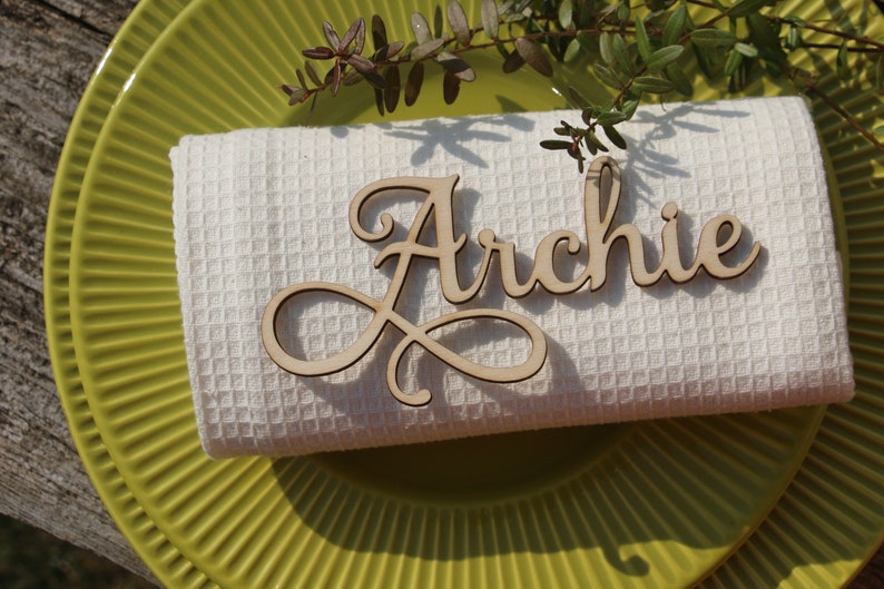 Puede incluir: Una servilleta de tela blanca con una tarjeta de nombre de madera que dice "Archie" en escritura cursiva. La servilleta est&aacute; encima de un plato verde.