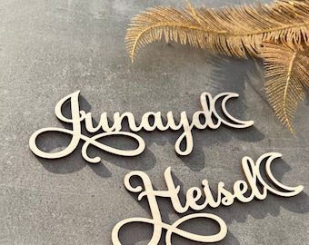 Op de afbeelding: Twee houten naamuitsparingen, "Junayd" en "Heisel", in een cursief lettertype. De namen zijn lichtbruin en liggen op een grijze ondergrond. Op de achtergrond een goudkleurige veer.