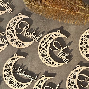 Puede incluir: Un conjunto de ocho adornos de madera en forma de media luna con intrincados diseños recortados. Cada adorno presenta un nombre diferente, incluyendo Zain, Rashid, Aisha, Muhammad, Omar, Fatima, Ibrahim y Zaid.