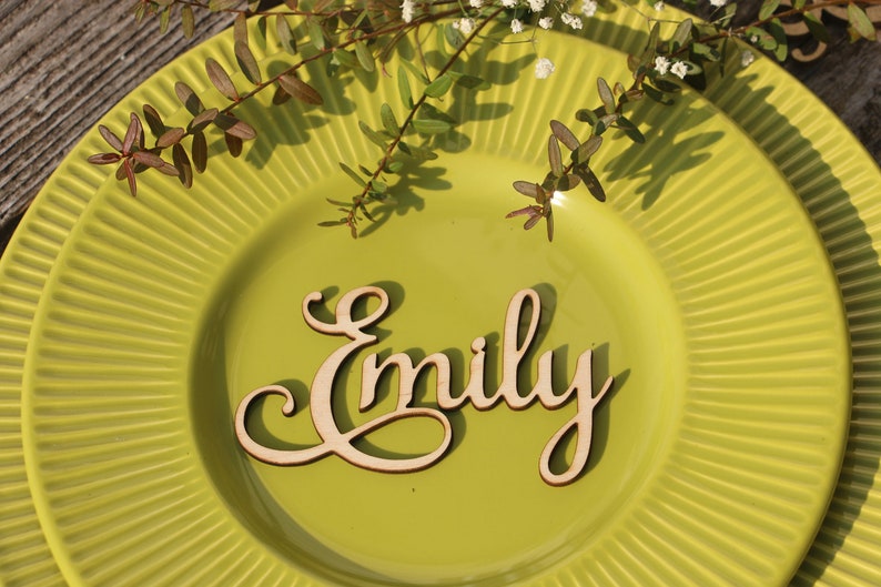 Puede incluir: Un tarjetero de madera con el nombre "Emily" escrito en cursiva. El tarjetero est&aacute; en un plato verde.