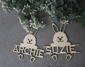 May include: Two wooden bunny-shaped name tags. Each bunny has a smiling face and holds a nameplate. One reads "ARCHIE", the other "SUZIE". The tags are light brown and sit on a gray surface, with white flowers and green leaves in the background.