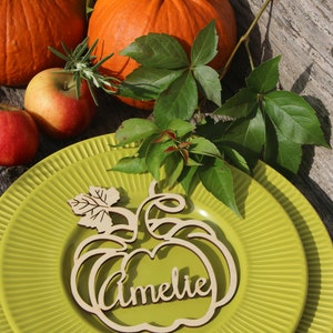Puede incluir: Una tarjeta de lugar de madera en forma de calabaza con el nombre "Amelie" escrito en ella. La tarjeta de lugar est&aacute; sobre un plato verde con una calabaza y hojas en el fondo.