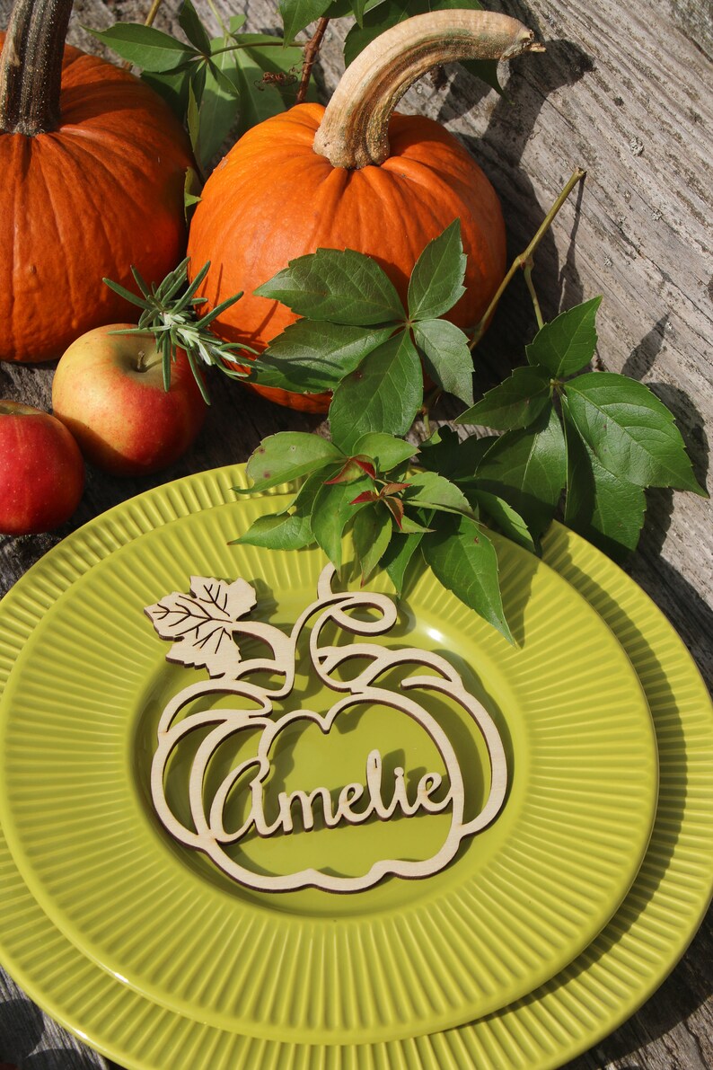 Puede incluir: Una tarjeta de lugar de madera en forma de calabaza con el nombre "Amelie" escrito en ella. La tarjeta de lugar est&aacute; sobre un plato verde con una calabaza y hojas en el fondo.