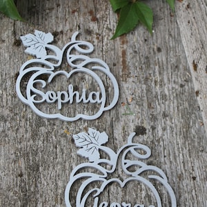 Puede incluir: Dos porta tarjetas de madera de color gris en forma de calabaza con los nombres "Sophia" y "George" escritos en ellos. Los porta tarjetas est&aacute;n sobre una superficie de madera con una hoja verde y una calabaza.