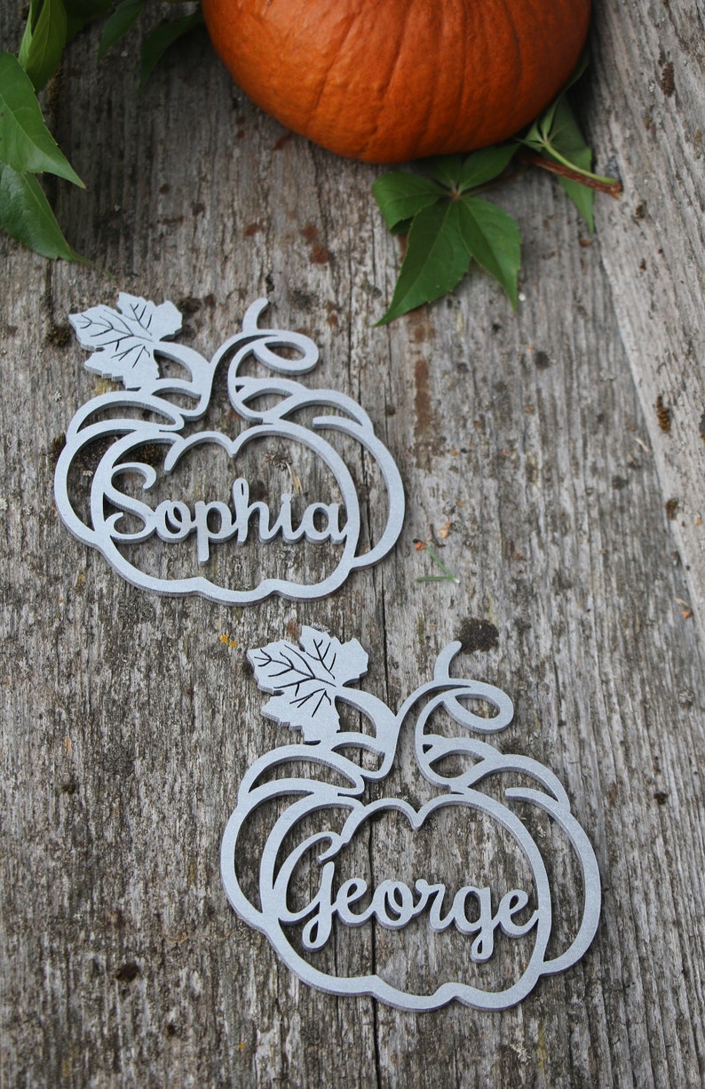 Puede incluir: Dos porta tarjetas de madera de color gris en forma de calabaza con los nombres "Sophia" y "George" escritos en ellos. Los porta tarjetas est&aacute;n sobre una superficie de madera con una hoja verde y una calabaza.