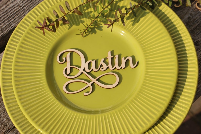 Puede incluir: Una tarjeta de lugar de madera con el nombre "Dustin" escrito en cursiva sobre un plato verde.
