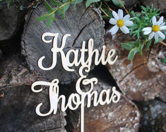 Puede incluir: Un letrero de madera blanco con los nombres "Kathie & Thomas" en escritura cursiva, mostrado sobre un fondo de troncos de madera apilados y margaritas blancas.