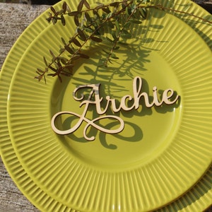 Puede incluir: Una tarjeta de lugar de madera con el nombre "Archie" escrito en cursiva sobre un plato verde con un dise&ntilde;o acanalado.