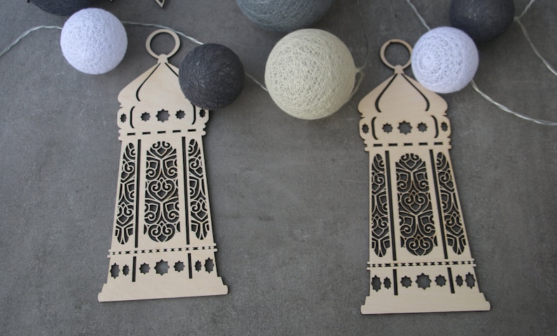 May include: Two wooden cutout lanterns with intricate designs, hanging from a string of white cotton balls. The lanterns are beige and have a dark brown intricate design.