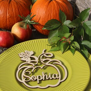 Puede incluir: Una tarjeta de lugar de calabaza de madera con el nombre "Sophia" tallado en ella. La tarjeta de lugar est&aacute; en un plato verde con un borde decorativo. Hay dos calabazas y manzanas en el fondo.