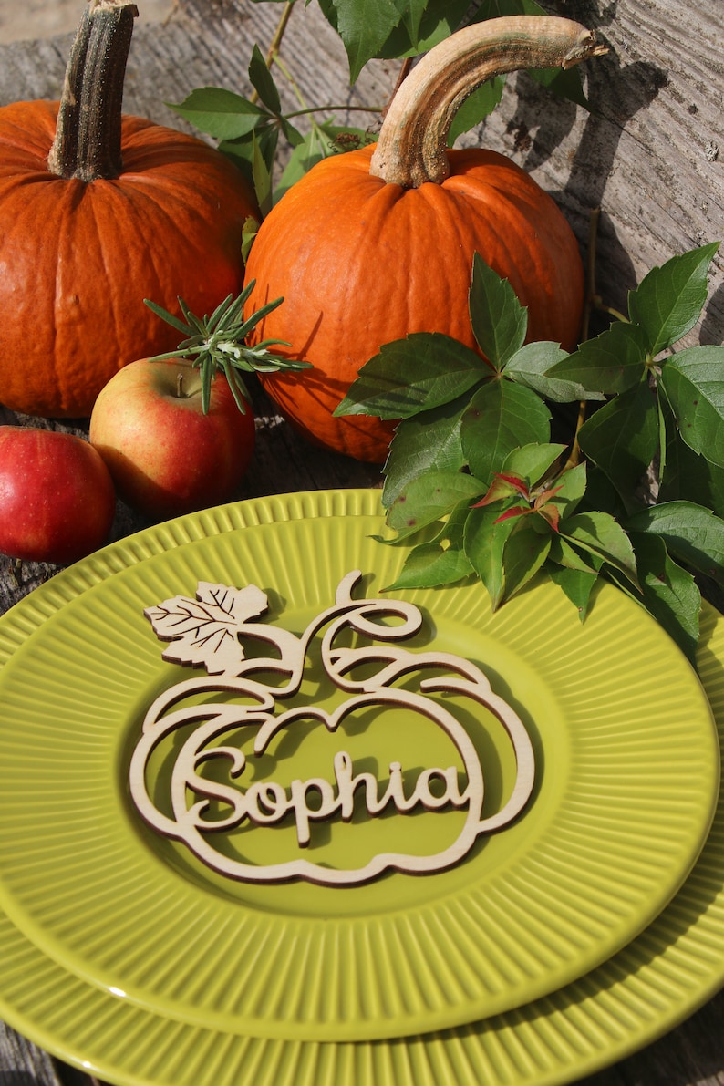 Puede incluir: Una tarjeta de lugar de calabaza de madera con el nombre "Sophia" tallado en ella. La tarjeta de lugar est&aacute; en un plato verde con un borde decorativo. Hay dos calabazas y manzanas en el fondo.