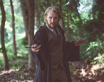 May include: A man wearing a black robe and brown vest stands in a forest, holding a wand in his right hand. He is looking directly at the camera.